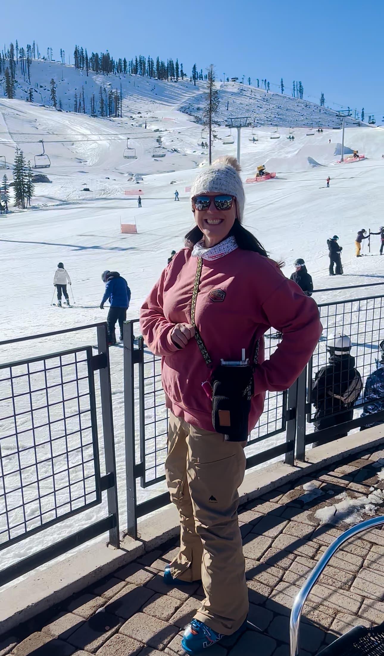 Nicola Banks, founder of Travola, smiling at a sunlit ski resort while carrying her Travola crossbody hydration purse hands-free across her shoulder.