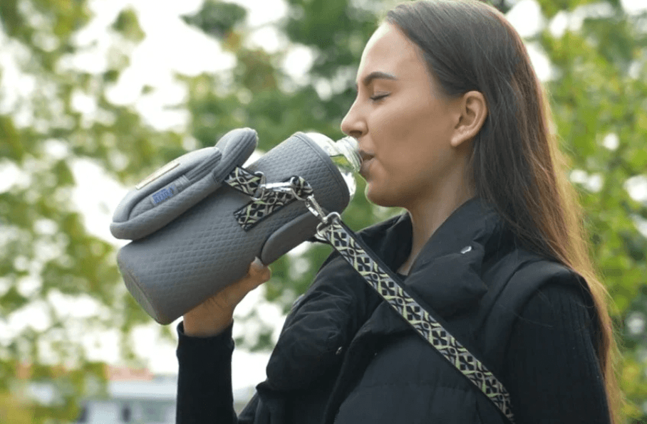 A woman sipping from her insulated bottle carried hands-free in the Travola Morning Fog grey crossbody hydration purse, with the woven adjustable strap across her shoulder.