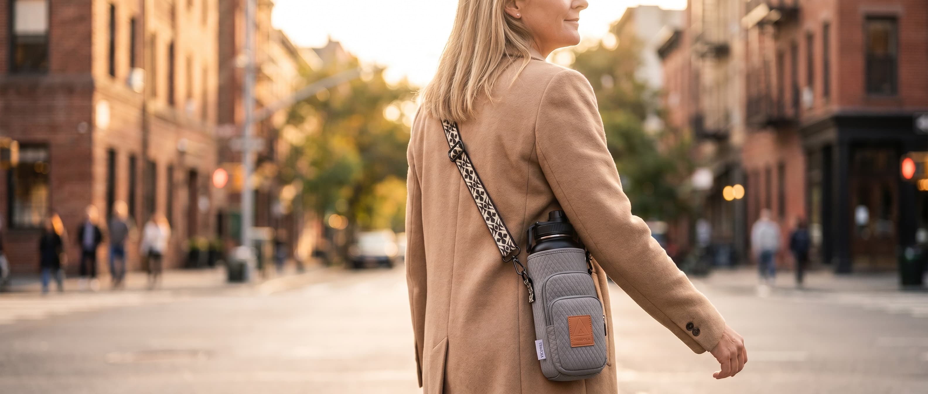 A woman walking a sunlit city street with the Travola neoprene water-bottle carrier worn crossbody, hands free.